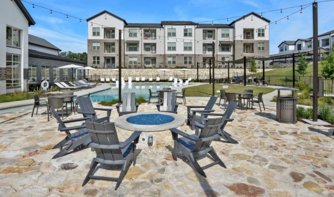 Outdoor patio area featuring a circular fire pit surrounded by gray lounge chairs, with tables and a pool in the background, and apartment buildings in view
