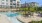 Outdoor pool area with lounge chairs, patio tables, and string lights, surrounded by apartment buildings and a landscaped grassy area in the background